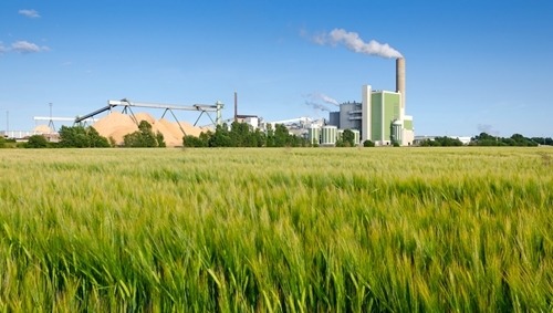 A lush green field with a large manufacturing facility in the background behind the field.