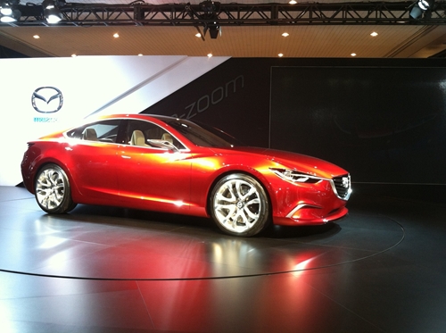 A brand new Mazda vehicle stands inside a large showroom with a big Mazda logo as the background.