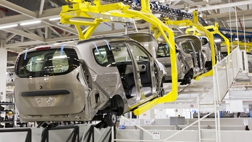 A series of robotic arms take the frame of a minivan from one end of a manufacturing facility to another.